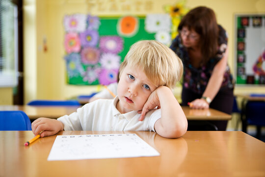 A tired young school boy resting his head in a classroom. He is looking bored or sleepy with his head on his hand while sitting at his desk with his teacher in the background. From a series of images. - Powered by Adobe