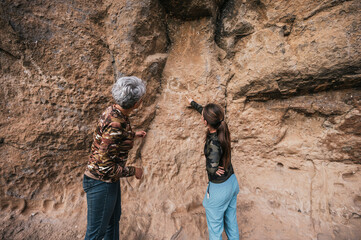 Mother and daughter dressed in hiking clothes, walking along a cliff observing cave paintings.	
