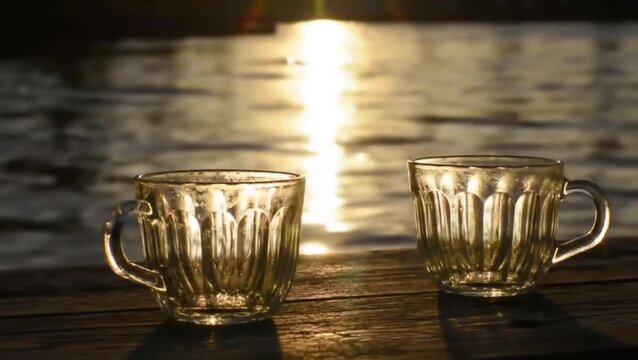 Two empty glass cups placed on a wooden surface with sunlight reflecting on the water in the background during golden hour. Still life concept