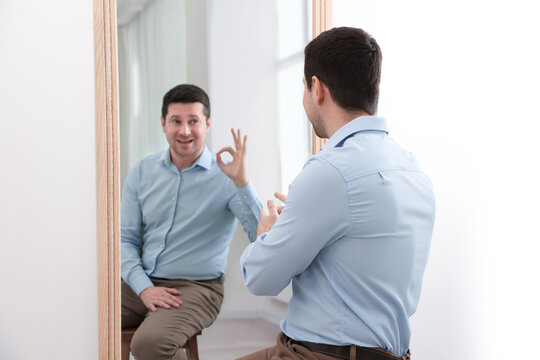 Man sitting in front of mirror where his reflection showing Ok gesture indoors. Mental health