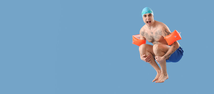 Handsome male swimmer with inflatable armbands doing cannonball jump on blue background