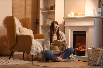 Young woman with plaid reading book near fireplace at home in evening