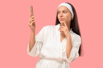 Thoughtful young woman in bathrobe and headband with mascaras on pink background