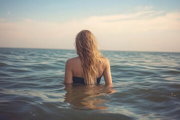 Obraz premium Woman with long blonde hair standing chest-deep in calm ocean water facing the horizon under a partly cloudy sky during daytime