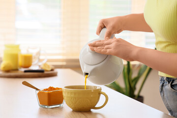 Woman pouring turmeric drink from teapot into cup at table in kitchen, closeup