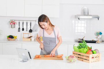Asian woman in a striped apron is seen chopping carrot on a wooden board in a bright, contemporary white kitchen, following a recipe on a tablet.