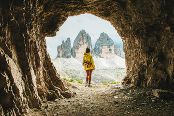 Explorer in Yellow Jacket Framed by Cave Opening with View of Tre Cime di Lavaredo Peaks