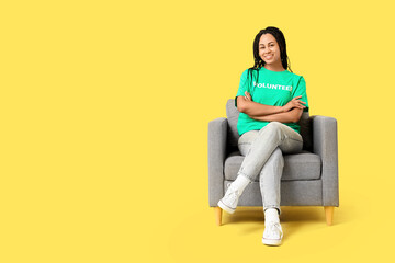 Young happy African-American female volunteer sitting on armchair against yellow background