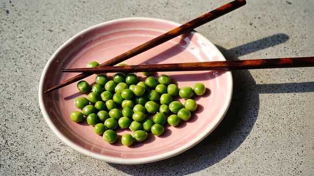 A plate of vibrant green peas with chopsticks resting on a pastel pink dish, set on a textured surface