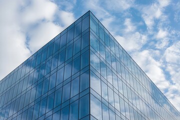 Obraz premium The sharp corner of a modern glass building is captured from a low angle, reflecting the blue sky and clouds in its sleek facade. 