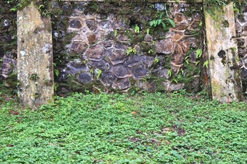 A retaining wall constructed of river stones and concrete columns as reinforcements that function to prevent landslides in an area with lush green grass growing around it