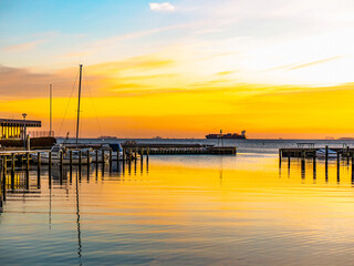 Obraz premium Sunrise over calm waters at Kastruphavn marina in Tårnby, Denmark, with boats, wooden piers, and a distant ship under a vibrant orange and blue sky.