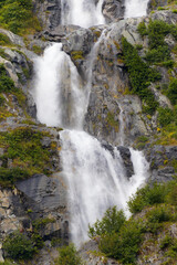 A powerful, multi-tiered waterfall cascades down a rocky Alaskan cliff, its white water contrasting sharply with the dark stone and lush green foliage growing on the steep face.