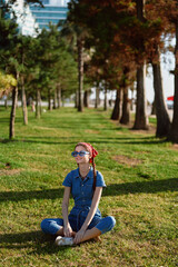 Fototapeta premium Lifestyle woman in modern boho-western denim street style sitting cross-legged on grass, wearing sunglasses and red headscarf, sunny outdoor park with trees and urban background.
