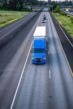 Bright blue big rig high cab semi truck transporting cargo in dry van semi trailer running on the morning straight highway road