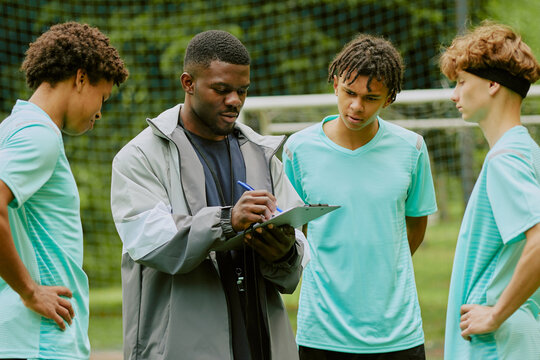 Black man coaching group of multiethnic teenage boys on soccer field, holding clipboard and explaining strategy while teenagers listening attentively during outdoor sports training