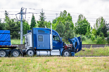 Broken blue big rig semi truck tractor with open hood and loaded flat bed semi trailer standing on...