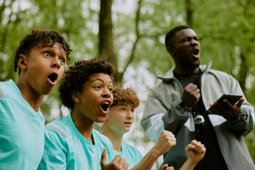 Group of multiethnic teenage boys cheering with excited expressions outdoors, Black man coach standing behind holding digital tablet and shouting encouragement during sports event
