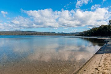 The crystal-clear waters of a tranquil lake, with mountains in the distance. Wallagoot Lake in Bournda National Park, South Coast NSW, Australia