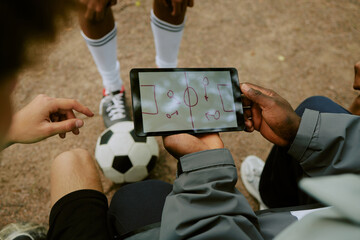 Caucasian teenager and Black man analyzing soccer strategy on digital tablet outdoors, soccer ball on ground, legs of teenager in sports uniform visible, teamwork and coaching concept