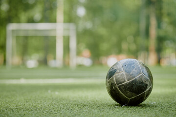 Old soccer ball resting on green artificial turf with blurred goalpost in background, outdoor sports field scene emphasizing worn equipment and recreational environment © Seventyfour