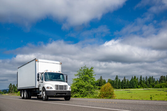 Compact middle duty day cab white rig semi truck transporting cargo in box trailer running on the local highway intersection