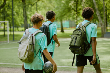 Fototapeta premium Three teenage boys, one Caucasian and two Black, walking together on outdoor soccer field carrying backpacks, one holding soccer ball, heading away from camera toward green park area