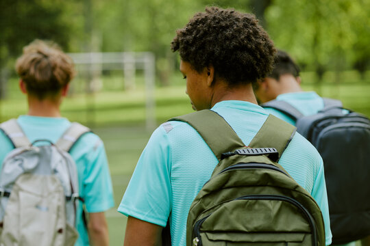 Group of teenage boys with backpacks walking outdoors on grassy field, multiethnic teenagers moving away from camera, casual clothing, blurred background with trees and soccer goal - Powered by Adobe