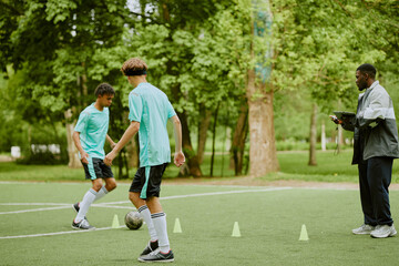 Two teenage boys, one Black and one Caucasian, dribbling soccer ball through cones on outdoor field while Black man coach observing during training session