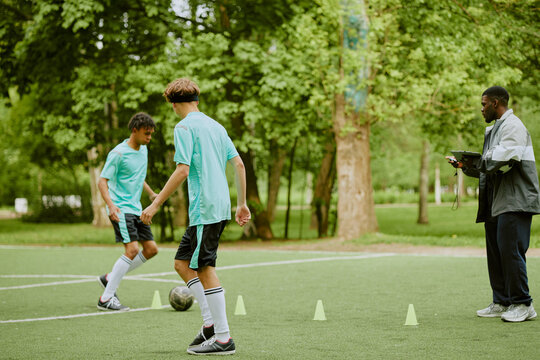 Two teenage boys, one Black and one Caucasian, dribbling soccer ball through cones on outdoor field while Black man coach observing during training session - Powered by Adobe