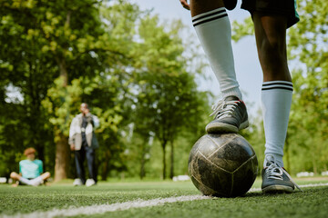 Teenage boy standing on soccer field placing foot on ball while two other teenagers sitting and standing in background on grass outdoors