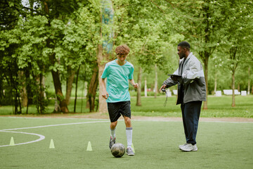 Teenage Caucasian boy dribbling soccer ball on outdoor field while Black male coach observing and giving instructions during training session in park