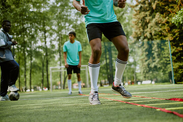 Teenage boys training on outdoor soccer field, Black teenager performing agility ladder drills in foreground while other teenage boys watching and preparing for soccer practice