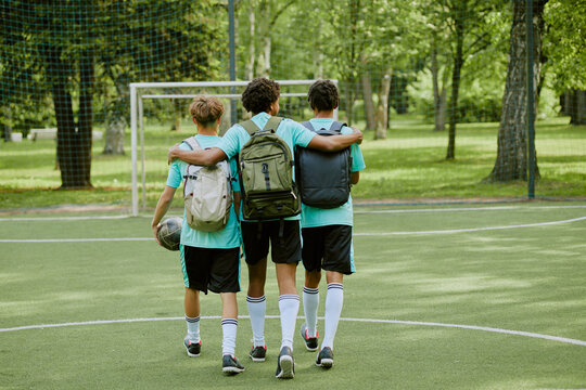 Three teenage boys, two Black and one Caucasian, walking together on outdoor soccer field with arms around each other, carrying backpacks, one holding soccer ball, viewed from behind