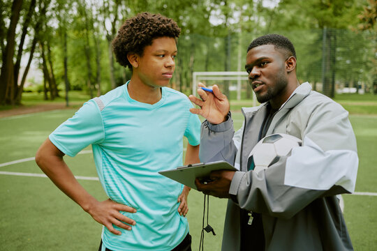 Teenager listening to Black male coach explaining strategy while holding soccer ball and clipboard on outdoor sports field, both standing close together during training session