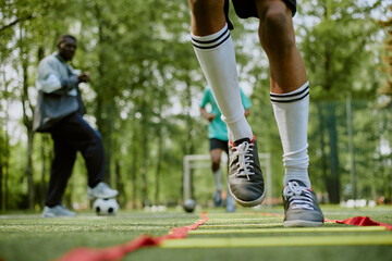 Teenager with athletic build performing agility ladder drill on outdoor soccer field while Black man coach observing in background, another teenager standing near soccer goal