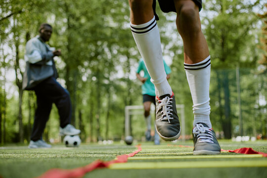 Teenager with athletic build performing agility ladder drill on outdoor soccer field while Black man coach observing in background, another teenager standing near soccer goal