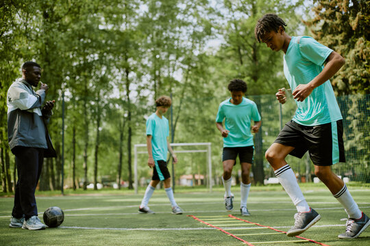 Group of teenage boys, including Black and Caucasian, training on outdoor soccer field, performing agility ladder drills while Black man coach observing and instructing near soccer ball