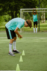Obraz premium Teenage Caucasian boy arranging training cones on outdoor soccer field while Black teenage boy standing in background near goalpost stretching arms above head