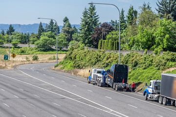 Powerful towing big rig semi truck picking up out of service broken black semi truck tractor standing on the highway road shoulder
