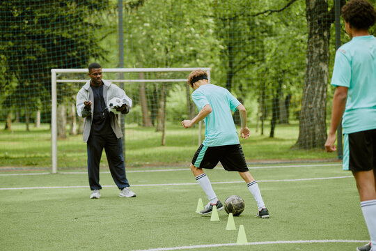 Teenage boys practicing soccer dribbling drills on outdoor field with Black man coach observing and holding soccer balls, green trees and netting visible in background - Powered by Adobe