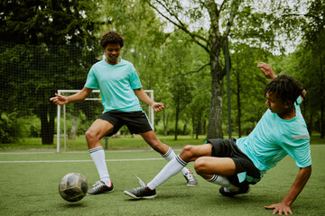 Two Black teenagers playing soccer on outdoor field, one teenager dribbling ball while other teenager sliding to tackle, green trees and netted goal visible in background