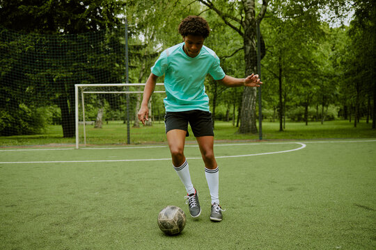 Teenage Black boy dribbling soccer ball on outdoor field, focusing on footwork and ball control, surrounded by green trees and empty goalpost in background during practice session