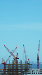Fototapeta premium Skyline view of a high-rise building under construction with multiple red cranes and steel framework, set against a clear blue sky in daylight.