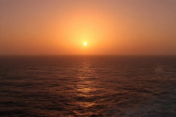 Sunset over a hazy ocean.  Warm, diffused light paints the sky and sea in soft oranges and reds.  A large sun dips below the horizon