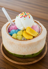 Coconut ice cream dessert served in a fresh coconut shell with colorful toppings, jackfruit, and a plastic spoon, sitting on a wooden plate.