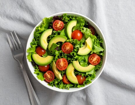 This image shows a bowl of fresh salad containing a mix of leafy greens, avocado slices, and halved cherry tomatoes. The leafy greens display a variety of textures and vibrant green shades. The avocad