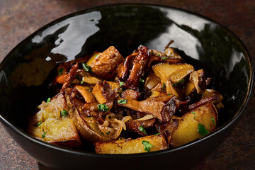 Fried potatoes with chanterelle mushrooms, rustic mood, closeup composition in dark ceramic bowl