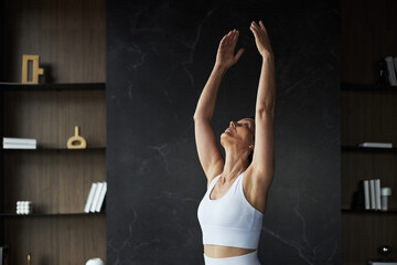Caucasian middle aged woman stretching arms upward during fitness routine, standing in modern interior, focusing on flexibility and wellness, wearing sportswear, looking up