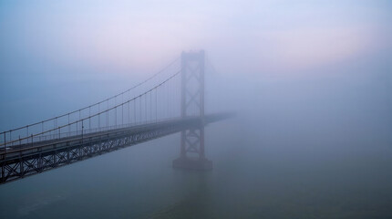 Fototapeta premium A suspension bridge is partially obscured by dense fog, creating a mystical and atmospheric scene over calm water.